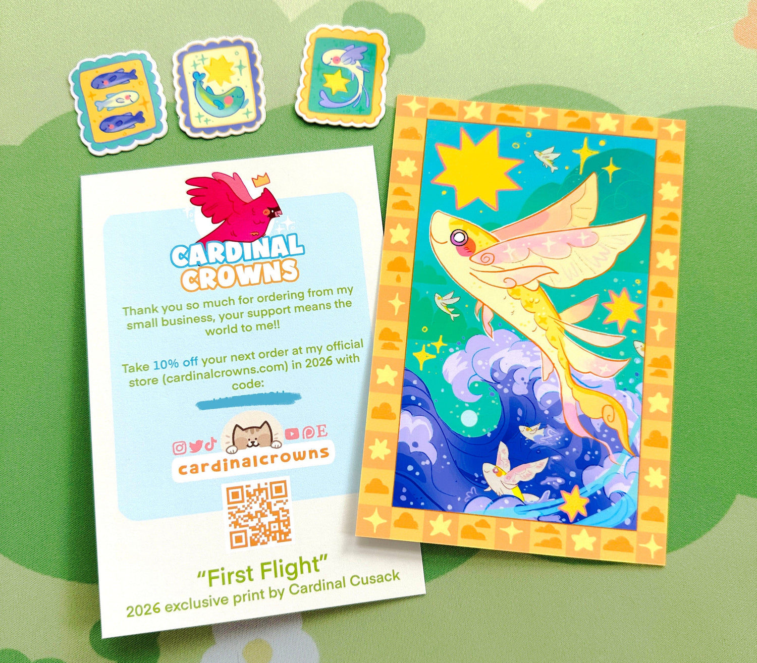 An aart print featuring a colorful fantasy illustration of a winged fish among stars and waves, shown alongside a white branded card and three small decorative stickers on a light green patterned surface.
